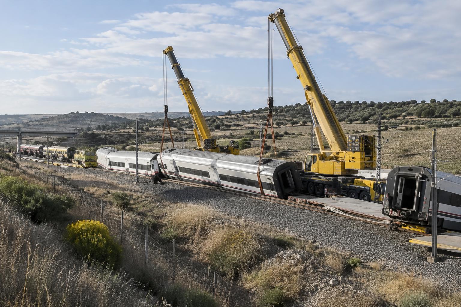 Zona de vías ferroviarias junto a un terraplén en un paisaje rural de Toledo, con grúas de gran tonelaje y vehículos de emergencia trabajando junto a vagones siniestrados, vista general sin víctimas visibles ni personas reconocibles.