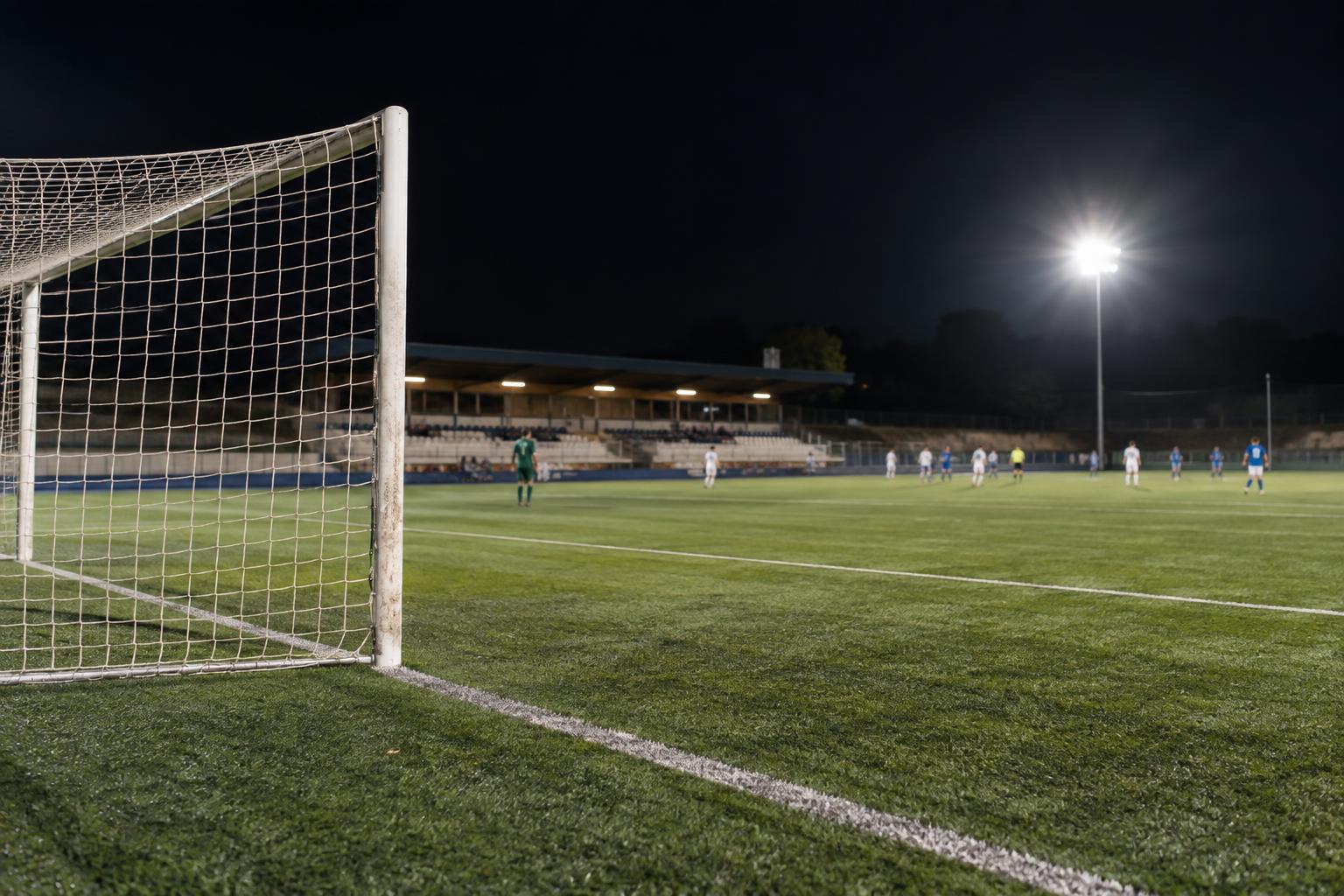 Campo de fútbol modesto español durante un partido nocturno, con césped iluminado por focos, una portería en primer plano y pequeñas gradas al fondo, sin personas reconocibles.