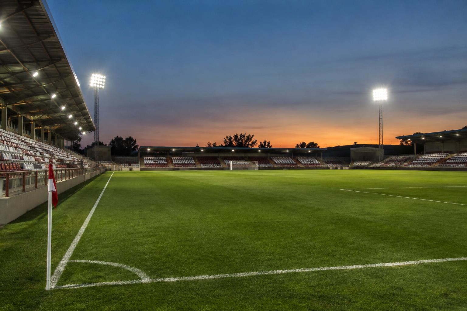 Vista realista de un estadio municipal de fútbol al anochecer, con césped iluminado por focos, líneas del terreno de juego y gradas modestas preparadas para un partido de Copa, sin personas reconocibles.