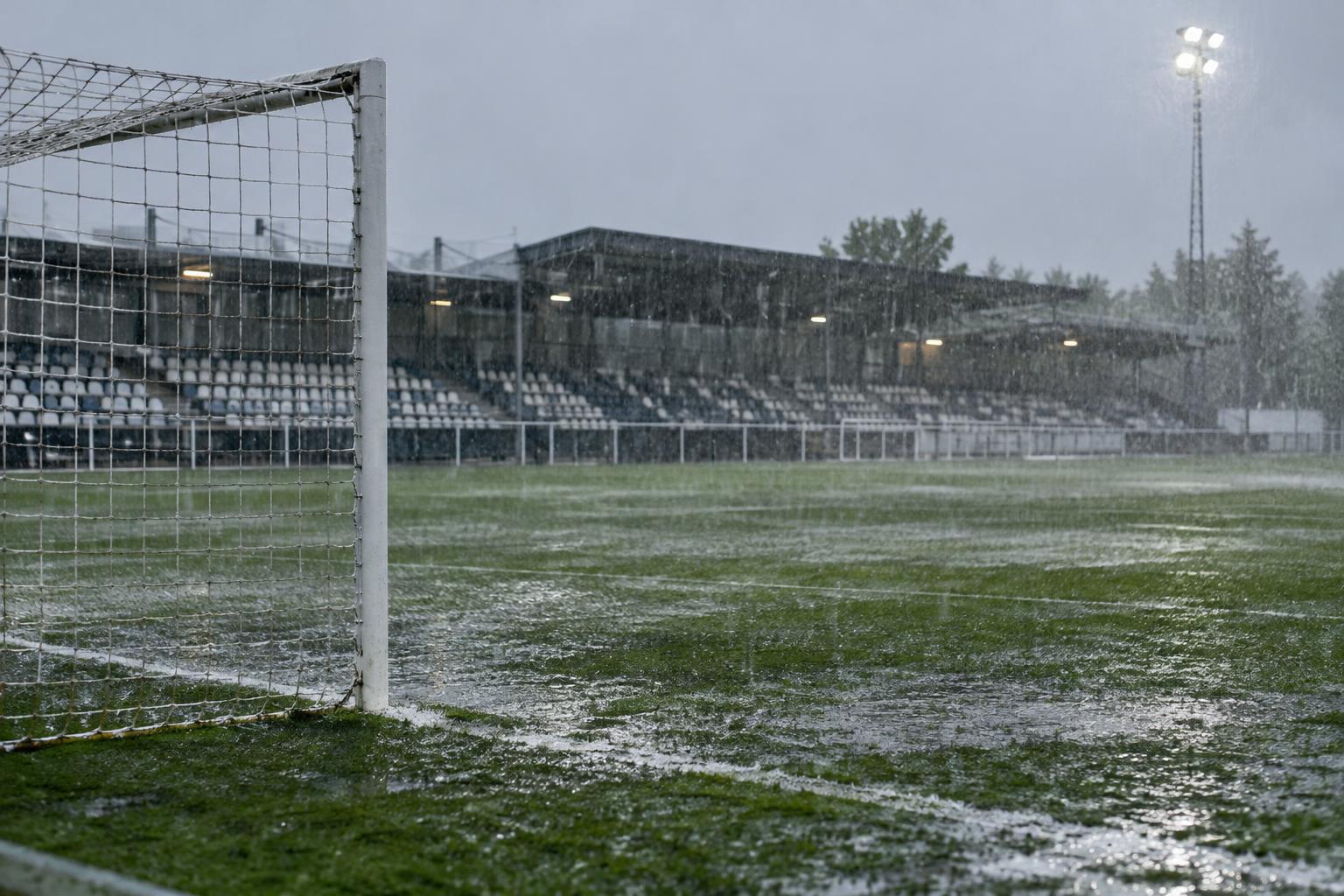 Campo de fútbol modesto bajo una lluvia intensa, con el césped mojado, una portería en primer plano y gradas cubiertas al fondo, sin personas reconocibles.