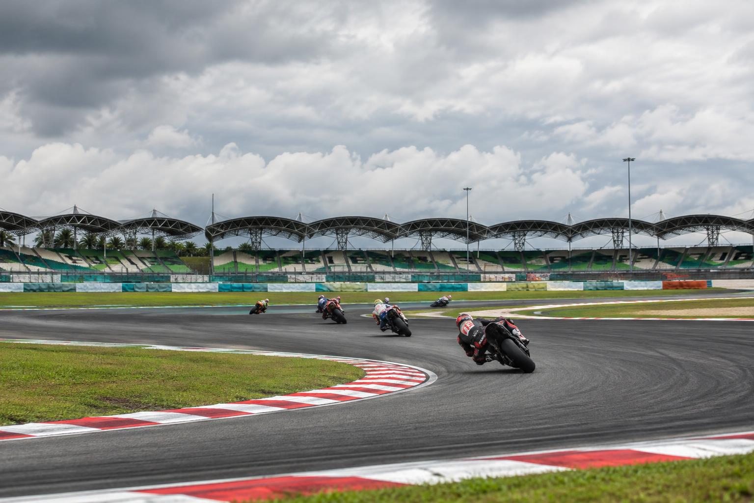 Vista realista de una curva del circuito de Sepang, con motos de competición tomadas desde lejos, asfalto oscuro, pianos de colores y tribunas al fondo bajo un cielo nublado, sin personas reconocibles.