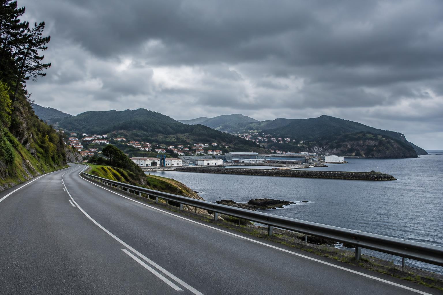 Carretera vacía junto a una pequeña localidad costera, montes boscosos al fondo y edificios portuarios bajo un cielo encapotado, encuadre panorámico realista con atmósfera inquietante, sin criaturas ni personas reconocibles.
