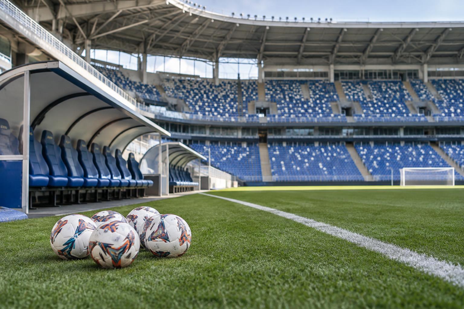 Vista realista a pie de césped de un estadio de fútbol con gradas blanquiazules vacías, el banquillo en primer plano y varios balones preparados junto a la línea de banda, sin personas reconocibles.