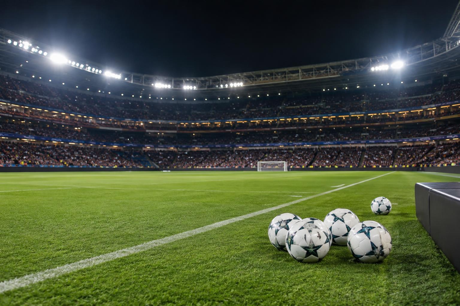 Vista realista del césped de un gran estadio de fútbol de noche, con las gradas concurridas, iluminación encendida y balones de calentamiento junto a la banda, sin personas reconocibles.