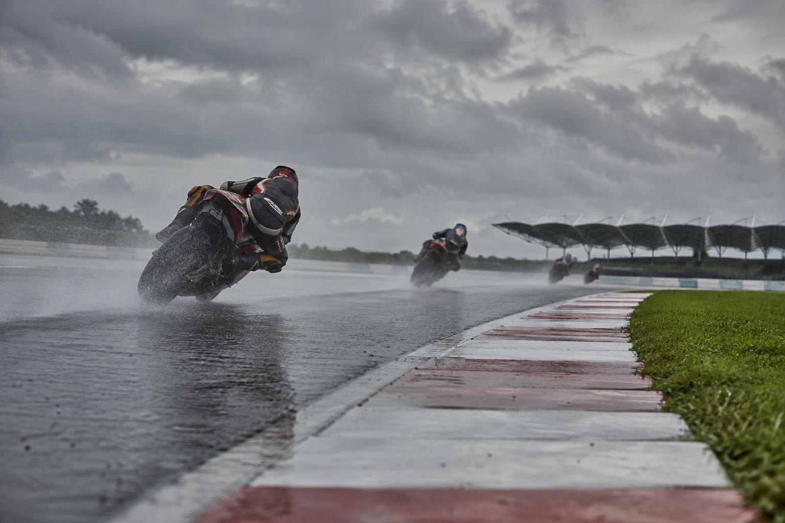Vista a ras de pista del circuito de Sepang, con asfalto parcialmente mojado, pianos de colores y varias motocicletas de competición al fondo bajo un cielo encapotado, sin rostros reconocibles.
