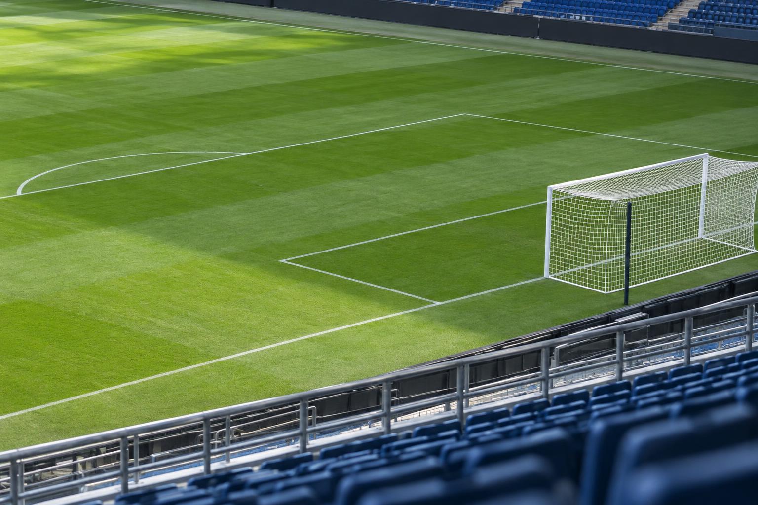 Vista realista de un campo de fútbol desde una grada lateral, con el césped, las líneas del área y una portería de red blanca, sin jugadores ni personas reconocibles.