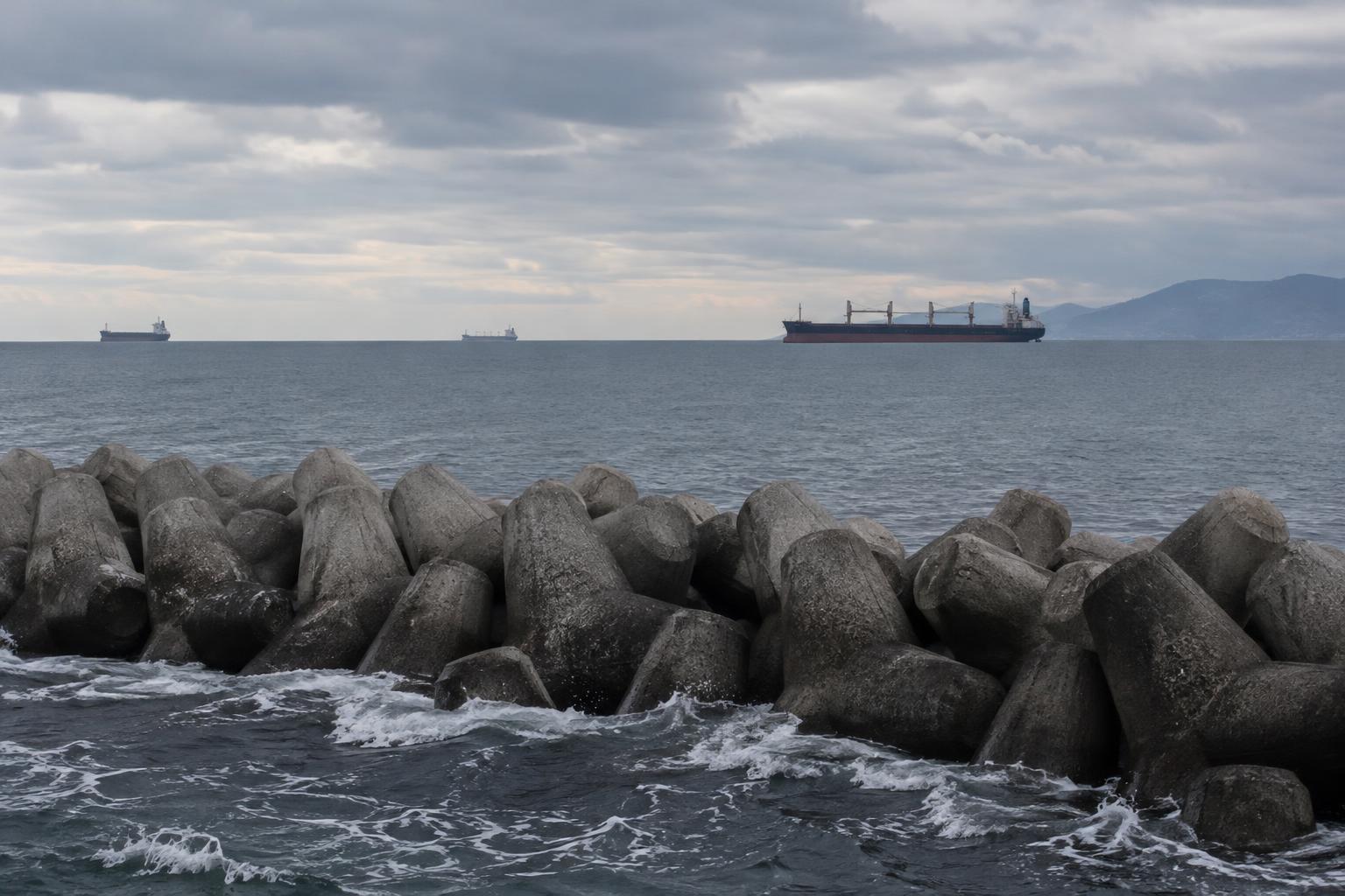 Vista realista de una costa del mar del Japón, con embarcaciones mercantes en el horizonte, un rompeolas en primer plano y cielo nublado, sin personas reconocibles ni explosiones.