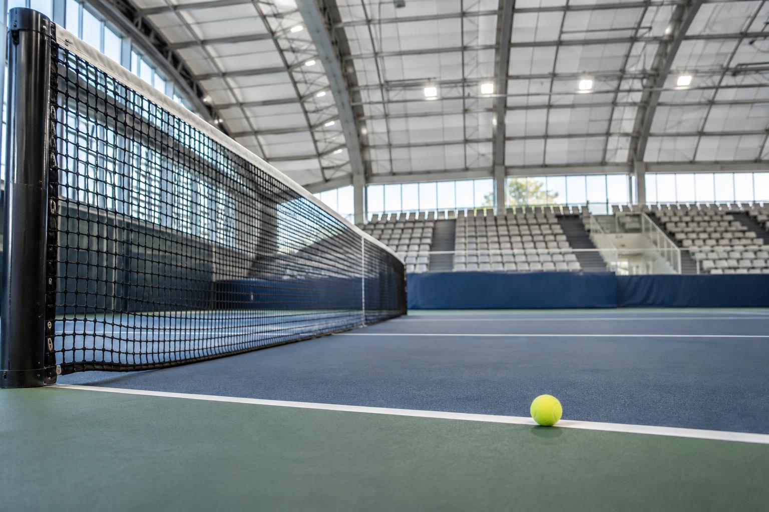Pista de tenis cubierta vista desde una esquina a ras de suelo, con la red en primer plano, una pelota junto a la línea de fondo y gradas iluminadas al fondo, sin personas reconocibles.