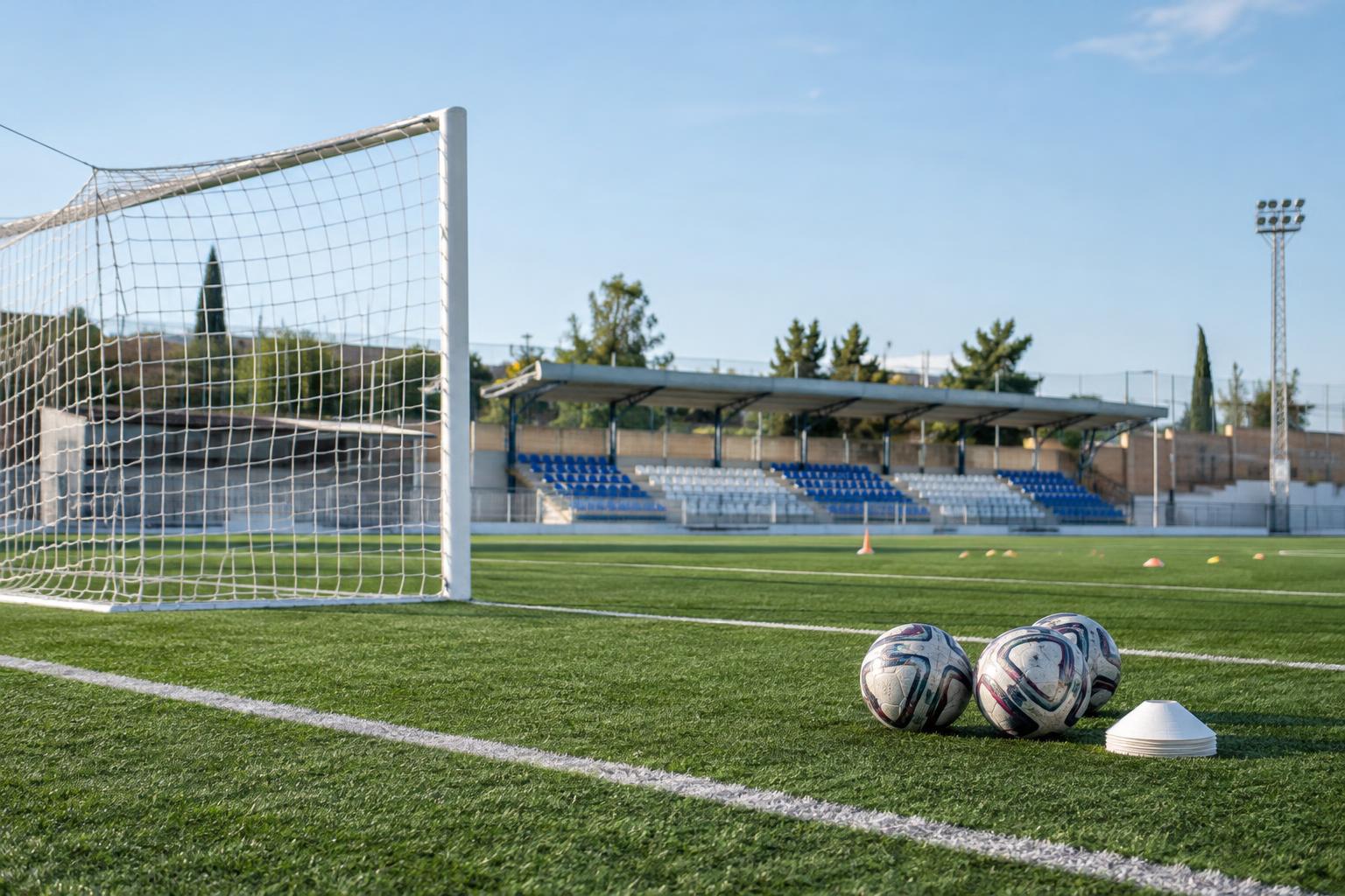 Campo de fútbol de césped sintético en una localidad española, con una portería, balones preparados para el calentamiento y una pequeña grada al fondo, sin personas reconocibles.