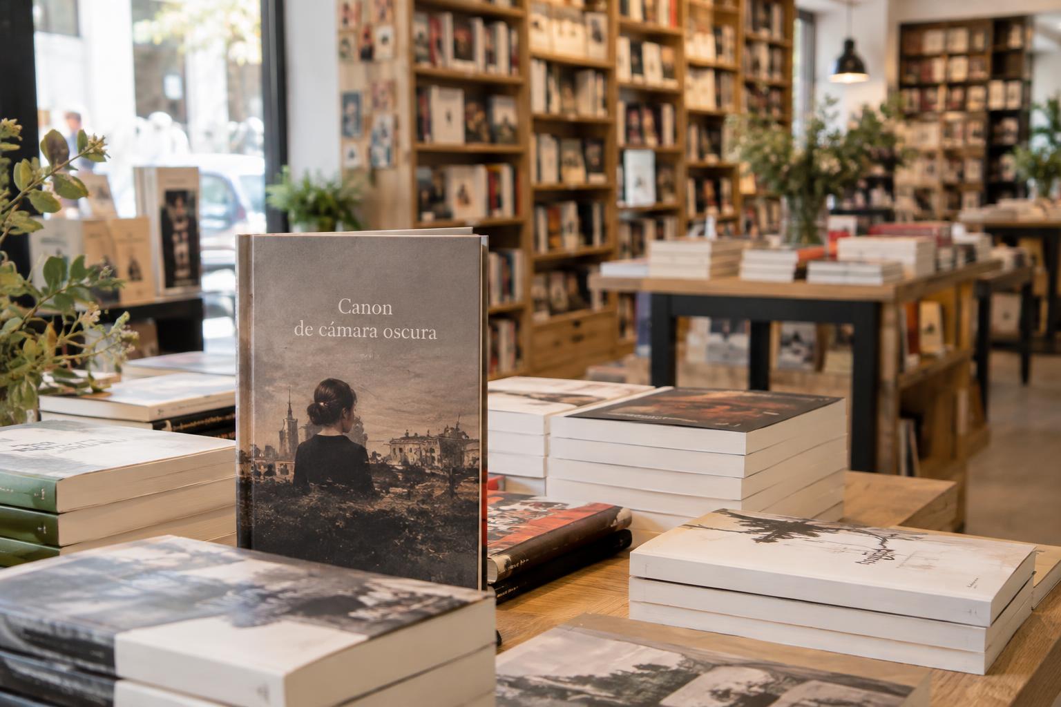 Interior de una librería de barrio, con mesas de narrativa española contemporánea, estanterías de madera y un ejemplar de Canon de cámara oscura en primer plano, sin personas reconocibles.