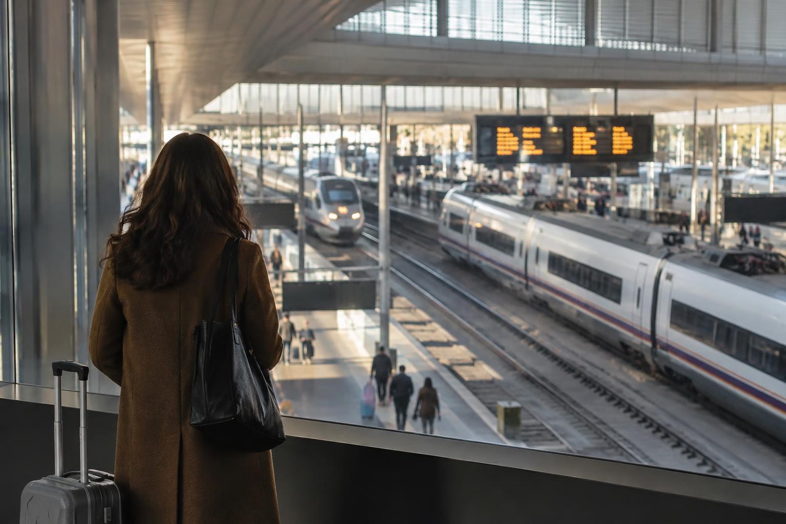 Interior realista de una estación ferroviaria española, con vías y andenes vistos a través de una cristalera, panel informativo al fondo y viajeros desenfocados sin rostros reconocibles.