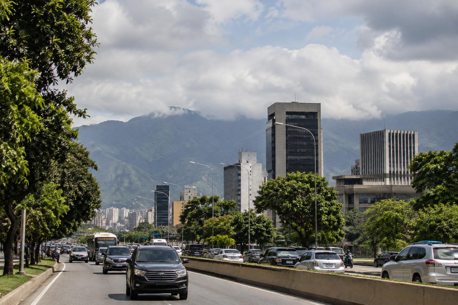 Vista realista de una avenida de Caracas con edificios institucionales al fondo, tráfico cotidiano y montañas bajo un cielo nublado, sin personas reconocibles.