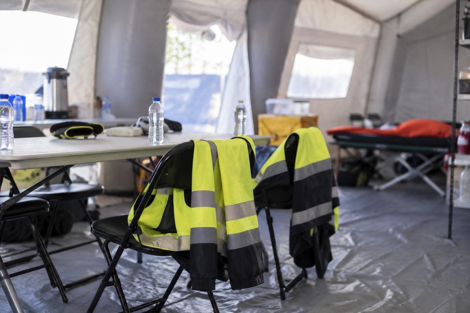 Interior de una zona provisional de descanso para equipos de emergencia, con sillas, botellas de agua y chaquetas reflectantes sobre los respaldos, luz natural y sin personas reconocibles.